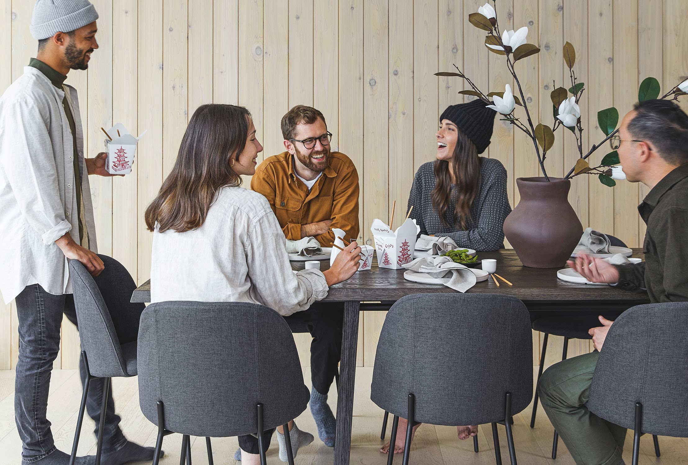 Cute people in toques gather around a dark wood table. Some are standing, most are seated in gray upholstered chairs.